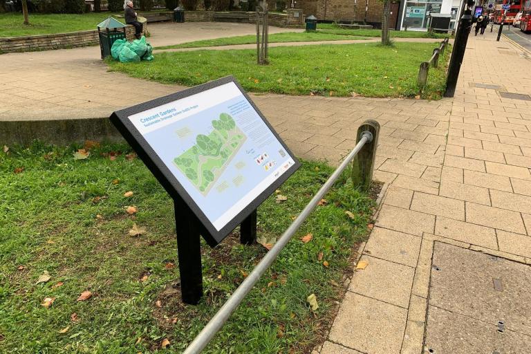 A sign at the edge of an area of grass. The sign has an illustration of the Crescent Gardens sustainable drainage project on it.